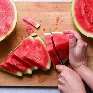 Jemand schneidet auf einem Brett eine Wassermelone. Symbolbild für Rezepte und den besten Trick, die Melone zu schneiden.