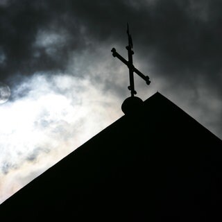 Im Gegenlicht und vor wolkenverhangenem Himmel ist die Kirchturmspitze des Doms in Osnarbrück mit Kreuz zu sehen.