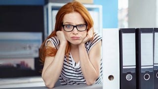 Junge Arbeitnehmerin sitzt mit frustriertem, fragendem Gesichtsausdruck bei der Arbeit und denkt über die Rechte und Konsequenzen von Arbeitsverweigerung nach. Neben ihr sind Aktenordner auf dem Tisch.
