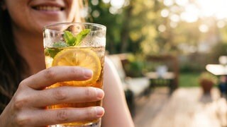 Jemand hält ein Glas mit Tee oder Limo im Sommer – sollte man diese Getränke bei Hitze wirklich nicht trinken?