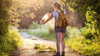 Eine Frau sprüht sich beim Wandern in der Natur ein Mittel gegen Mittel und Zecken auf den Arm. Welche empfiehlt Stiftung Warentest?