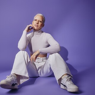 Stefanie Heinzmann sitzt weiß gekleidet vor einer blau-violetten Wand
