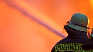 Symbolbild Feuerwehr beim Löschen zum Artikel über einen Autotransporter, der auf der A6 bei Kaiserslautern brennt und eine Vollsperrung ausgelöst hat. Man sieht einen Feuerwehrmann mit Schlauch und im Hintergrund ein großes Feuer.