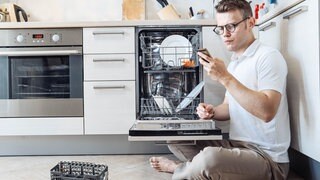 Ein Mann sitzt vor einer Spülmaschine und schaut auf dem Smartphone, was die Spülmaschinen-Symbole bedeuten