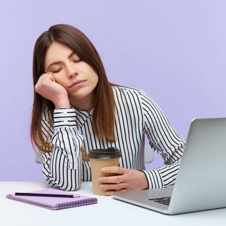 Eine schläfrige Frau sitzt im Büro am Laptop und ihr fallen die Augen zu. Ein Mittagschlaf bei der Arbeit kann gesund sein.