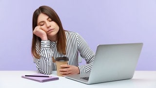 Eine schläfrige Frau sitzt im Büro am Laptop und ihr fallen die Augen zu. Ein Mittagschlaf bei der Arbeit kann gesund sein.