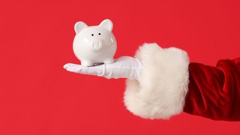Hand von einem Weihnachtsmann mit einem weißen Sparschwein als Symbol für Spenden in der Weihnachtszeit und welche Tipps man beachten sollte, um richtig zu spenden. 