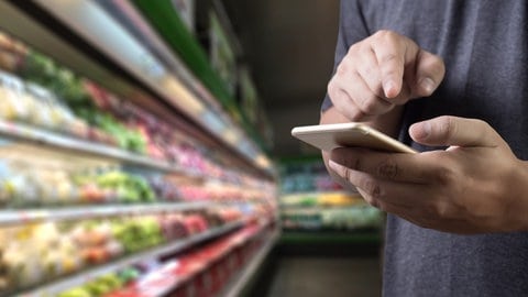 Männliche Hände tippen auf einem handy rum. Im Hintergrund die Kühltheke eines Supermarkts