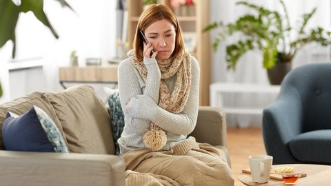 Eine Frau ist krank und möchte sich telefonisch vom Arzt krankschreiben lassen. So kann sie ihrem Arbeitgeber mitteilen, dass sie krank ist. 