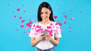 Eine junge Frau schaut auf ihr Handy und freut sich über Whatsapp-Sprüche und -Grüße zum Valentinstag. 