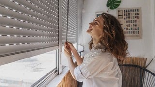 Junge Frau öffnet die Rolläden, aber sollte man sie bei Hitze ganz schließen sein oder nur halb?